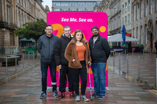 Group of four See Me volunteers stand in front of a pink banner reading 'See Me. See Us.'